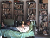 A man reclines while reading on a daybed draped with green linens. Behind him are four bookcases filled with vinyl records.
