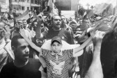 A young boy with his arms outstretched leans against a wall. The wall is covered with a larger than life photograph of a crowd of people holding flags and signs, as in a protest or a rally.