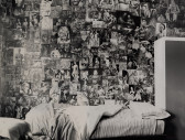 Black and white image of an unmade bed under a wall covered with small images of people and idols.