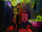 A figure wearing a trenchcoat stands in a psychadelically lit bathroom while smoking a cigarette.