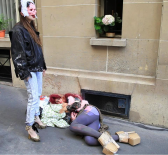 A woman in a mask smoking a cigarette standing over a person lying on blankets on the street wearing cardboard heels.