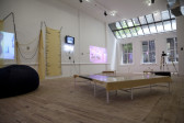 A gallery with an L-shaped bench covered in fabric, plus several video projections and artworks adorning the walls.