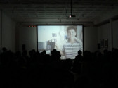 A crowd of people seated in a dark room, facing a screen. On screen, there is an image of a person wearing a striped shirt, holding a towel around their neck.