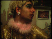 A person is seen in profile, speaking to someone out of frame. They are wearing a glittery gold top with a pink fur neckline and beaded necklaces of different colors. They have large flower-shaped earrings and colorful makeup, including blue, shiny eyeshadow. Their dark hair appears to be covered by a bleached wig.