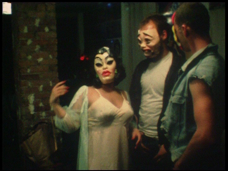 Three people wearing facemasks. One is dressed in an ultra-feminine mask, nightgown and open robe, facing the camera. The other two are facing the first figure, dressed casually in t-shirts and a denim vest.