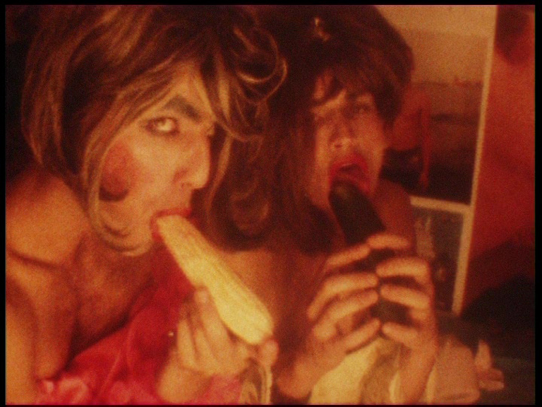 Two people with wigs and highly made up faces. The front figure is holding a cob of corn with the end in their mouth, looking suggestively at the camera. Behind them, a second figure holds a small eggplant with the end in their open mouth.