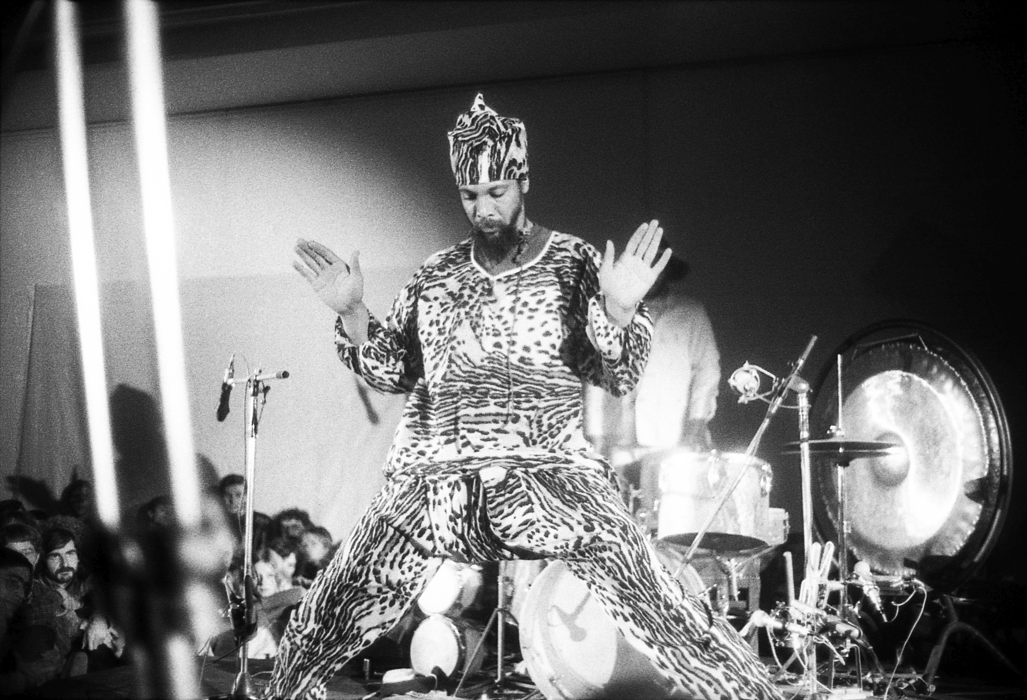 A black and white photograph of a man in a leopard print costume standing center stage with his hands up and palms open facing toward the viewer. He stands with his legs outstretched and behind him rests a drum set which sits next to a gong. Microphones are positioned near his body, one on each side.