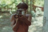 A shirtless man holding a film camera outdoors.