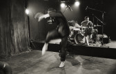 Black and white photograph of a performance. In the foreground, a blurred barefoot person in a suit swiftly raises their arms and right leg, curving in a halfmoon shape. In the background a drummer sitting at a drumset is visible.