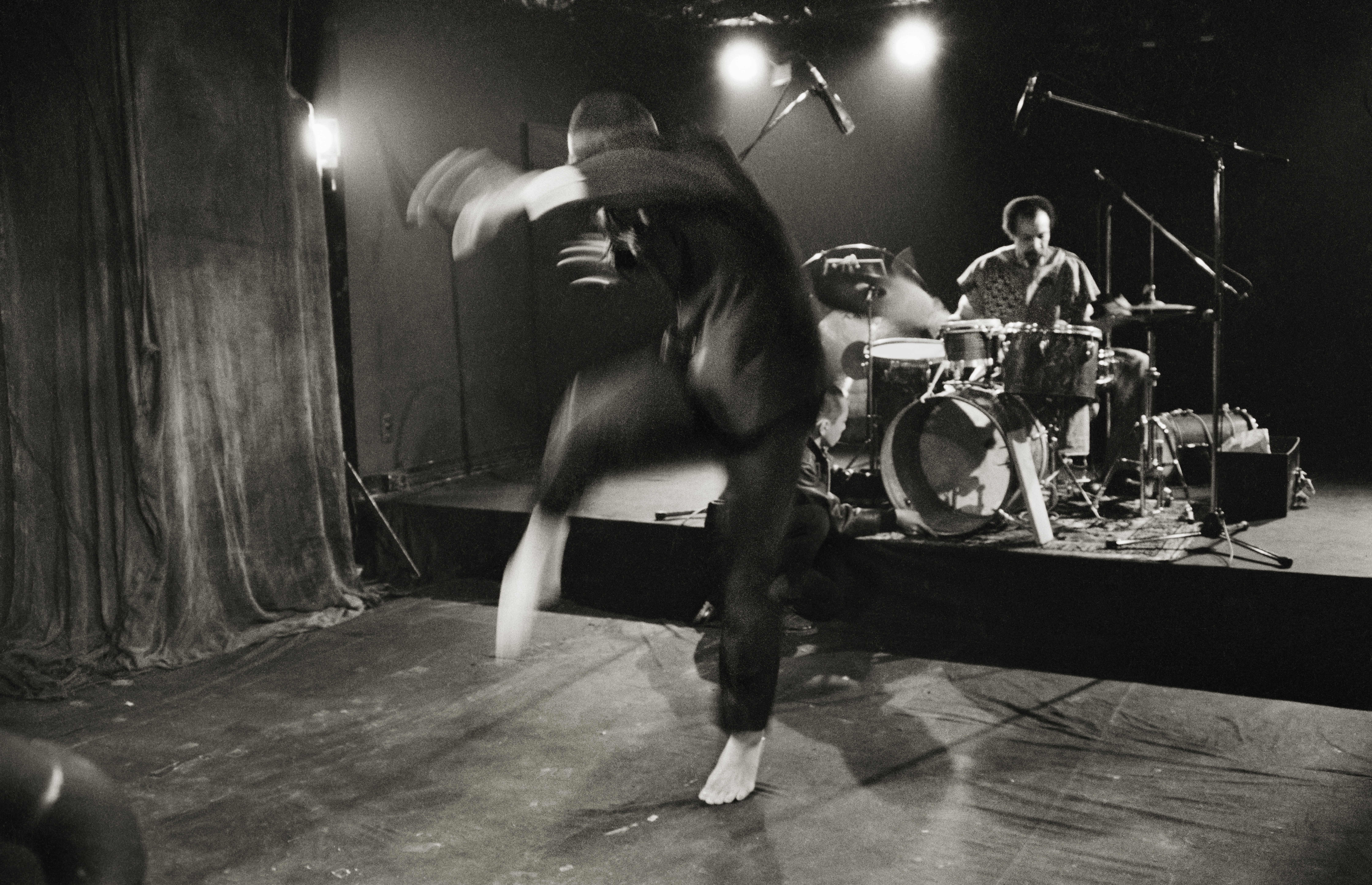 Black and white photograph of a performance. In the foreground, a blurred barefoot person in a suit swiftly raises their arms and right leg, curving in a halfmoon shape. In the background a drummer sitting at a drumset is visible.
