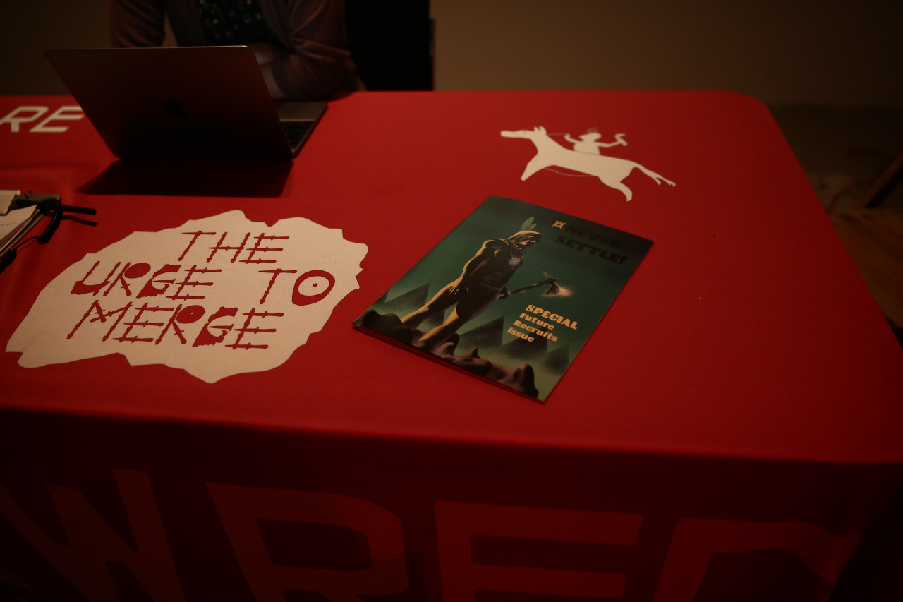 A table covered with a red tablecloth. The tablecloth is decorated with two white images: a person riding a horse and stylized text reading "THE URGE TO MERGE." Lying on the table, there is a magazine with a green cover with a figure holding an axe. The cover text reads, "NEVER SETTLE" and "SPECIAL Future Recruits Issue".