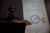 A person stands at a microphone reading off sheets of paper. In the background, there is a slightly blurry image of a book cover: 