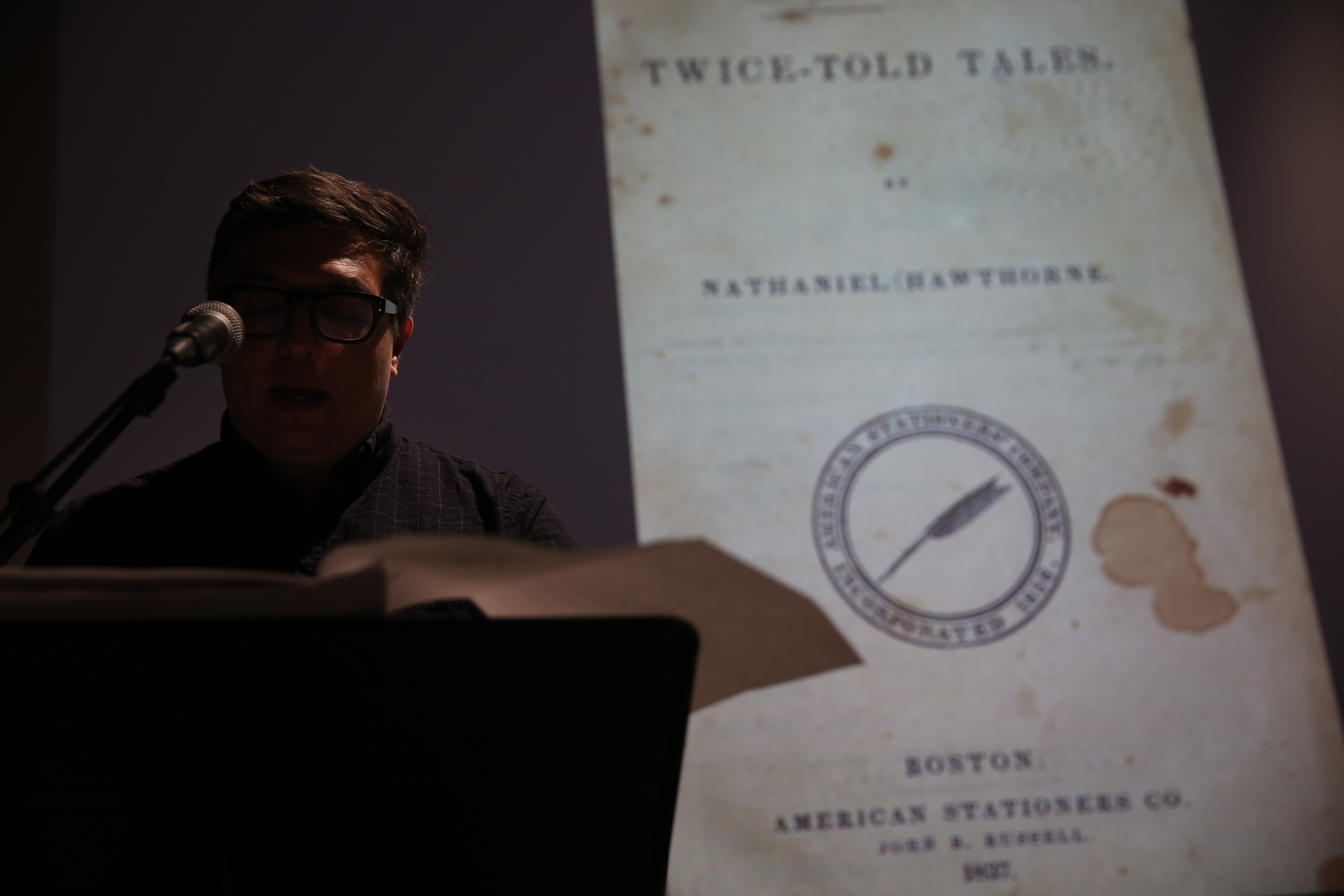 A person stands at a microphone reading off sheets of paper. In the background, there is a slightly blurry image of a book cover: 