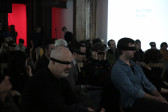 A crowd of people sitting in a dimly lit room. Most have their eyes obscured by black blindfolds. In the background, a white projection with black text just visible reads, 