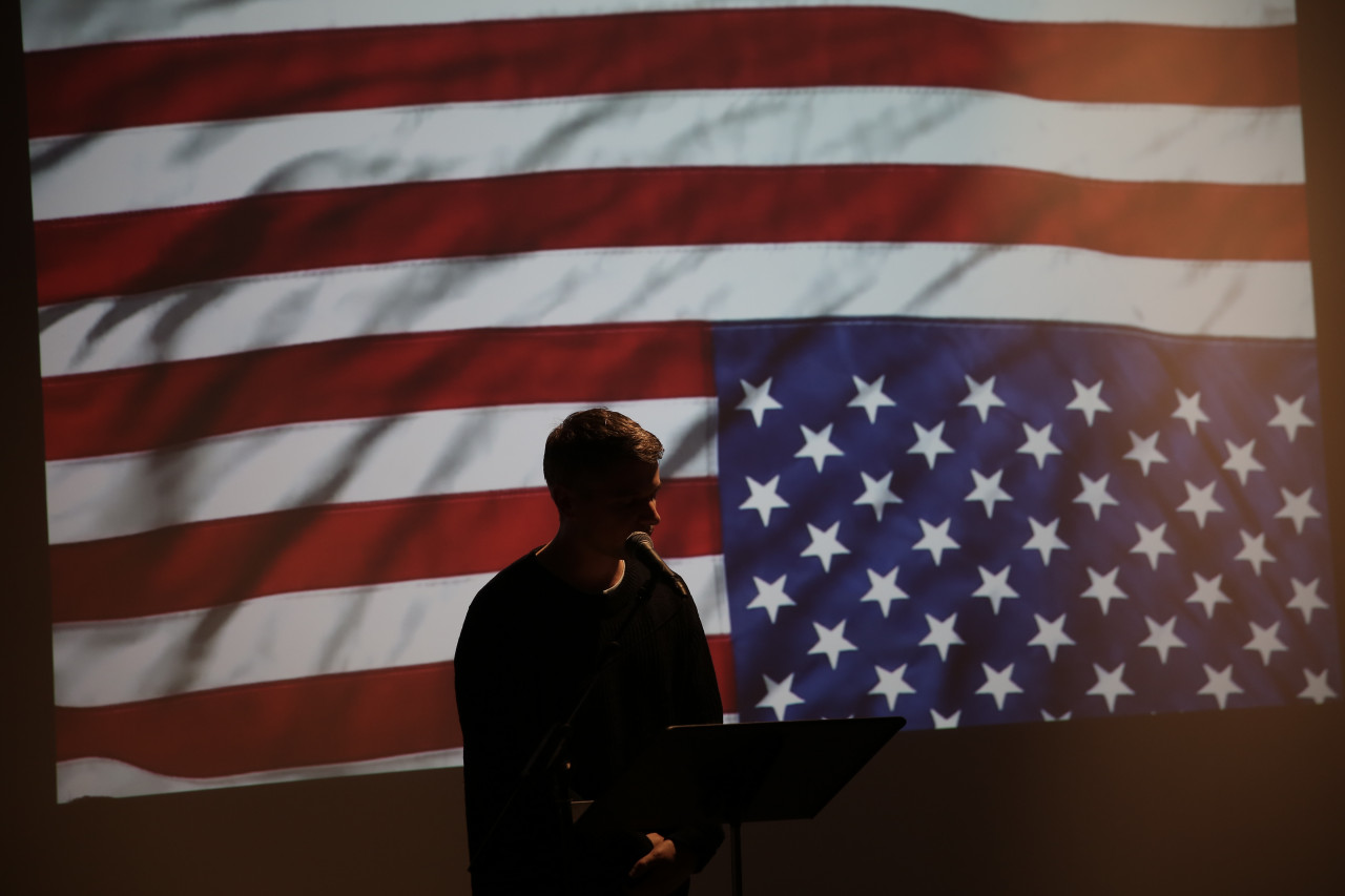 A silhouetted figure of a person speaking into a microphone. They are backlit by an image of an upside down American flag.