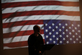 A silhouetted figure of a person speaking into a microphone. They are backlit by an image of an upside down American flag.