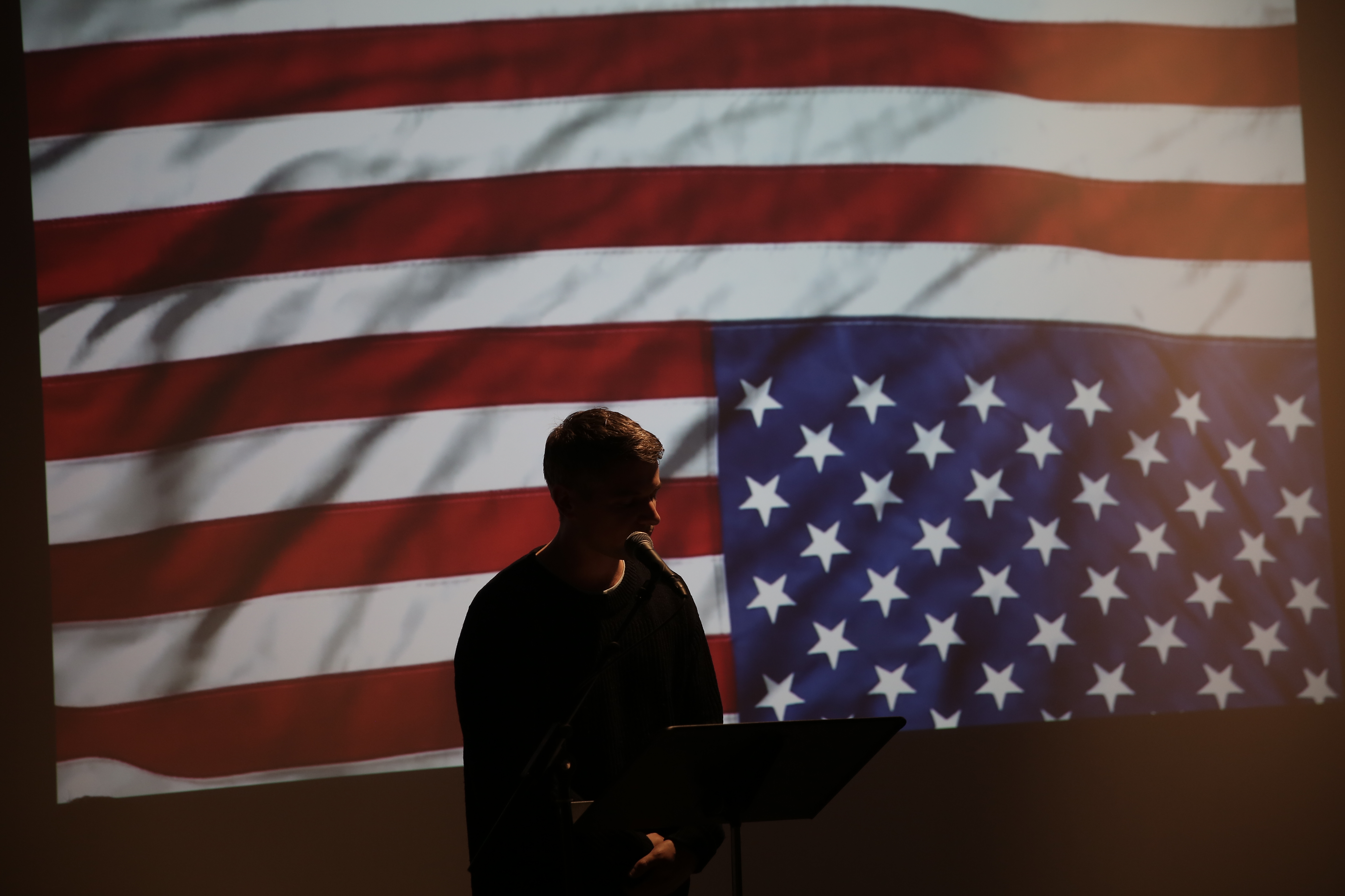 A silhouetted figure of a person speaking into a microphone. They are backlit by an image of an upside down American flag.