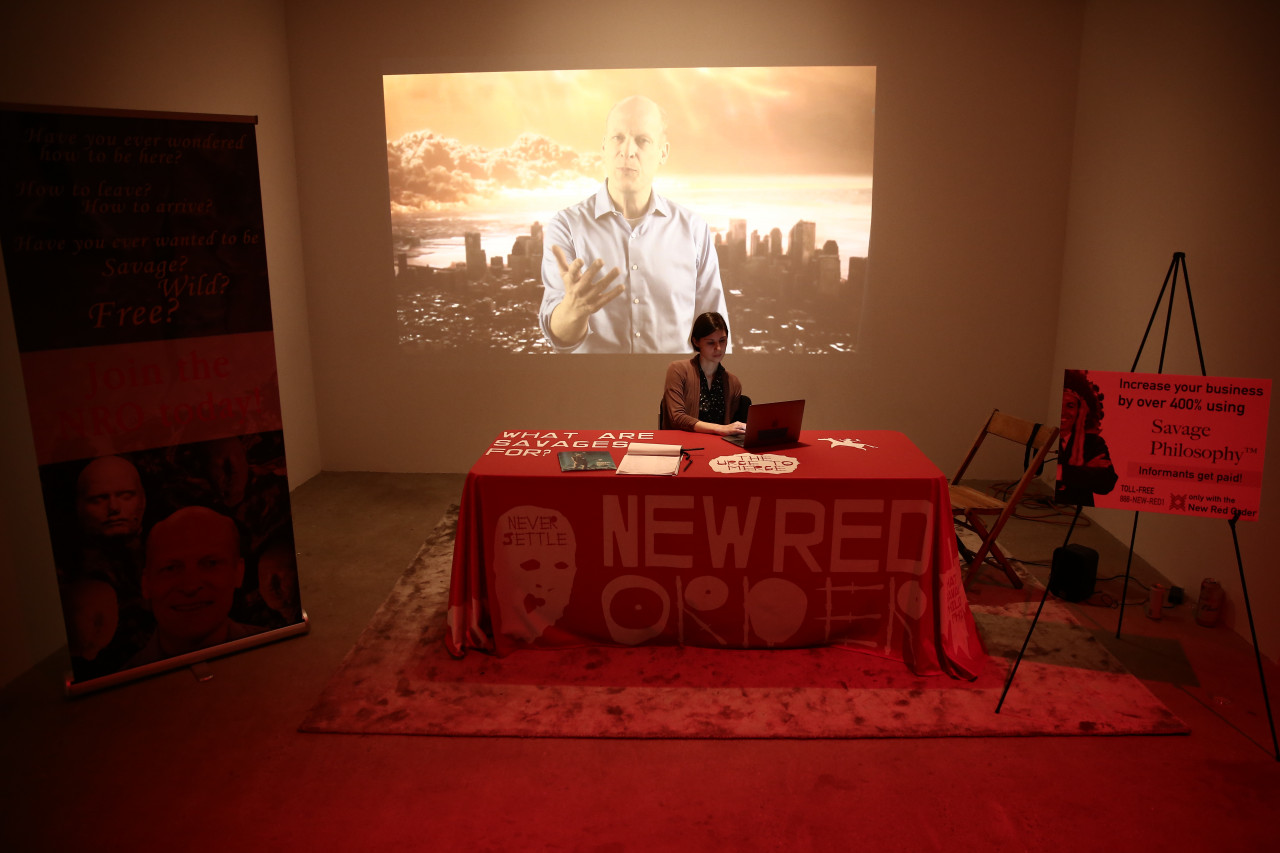 A wide shot of a person sitting at a table with a computer. The table is covered in a red tablecloth with white lettering that reads, "WHAT ARE SAVAGES FOR?" and "THE URGE TO MERGE." Lighter red, stylized text reads "NEW RED ORDER" and "NEVER SETTLE." Behind the table, a projected image shows a man with one hand outstretched superimposed on a city skyline. On either side of the table are informational posters with printed text.