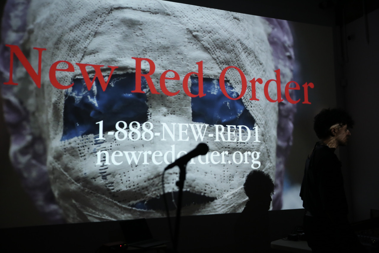 A figure and a microphone are silhouetted in front of an image projection. The image shows a face obscured with white plaster, overlaid with red text reading “New Red Order” and white text reading “1-888-NEW- RED1 newredorder.org."