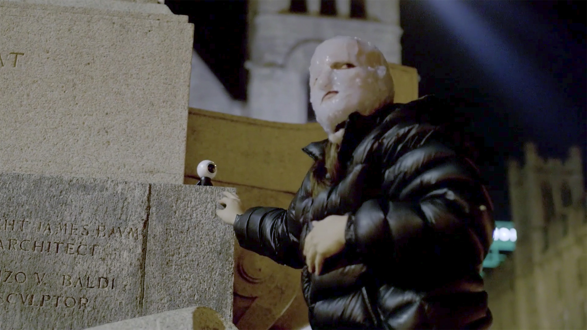 A figure wearing a black, puffy coat and a white, waxy mask that covers their entire head stands beside a stone structure with an illegible inscription. Their hand rests on the stone next to a small, spherical surveillance camera as they look off into the distance.