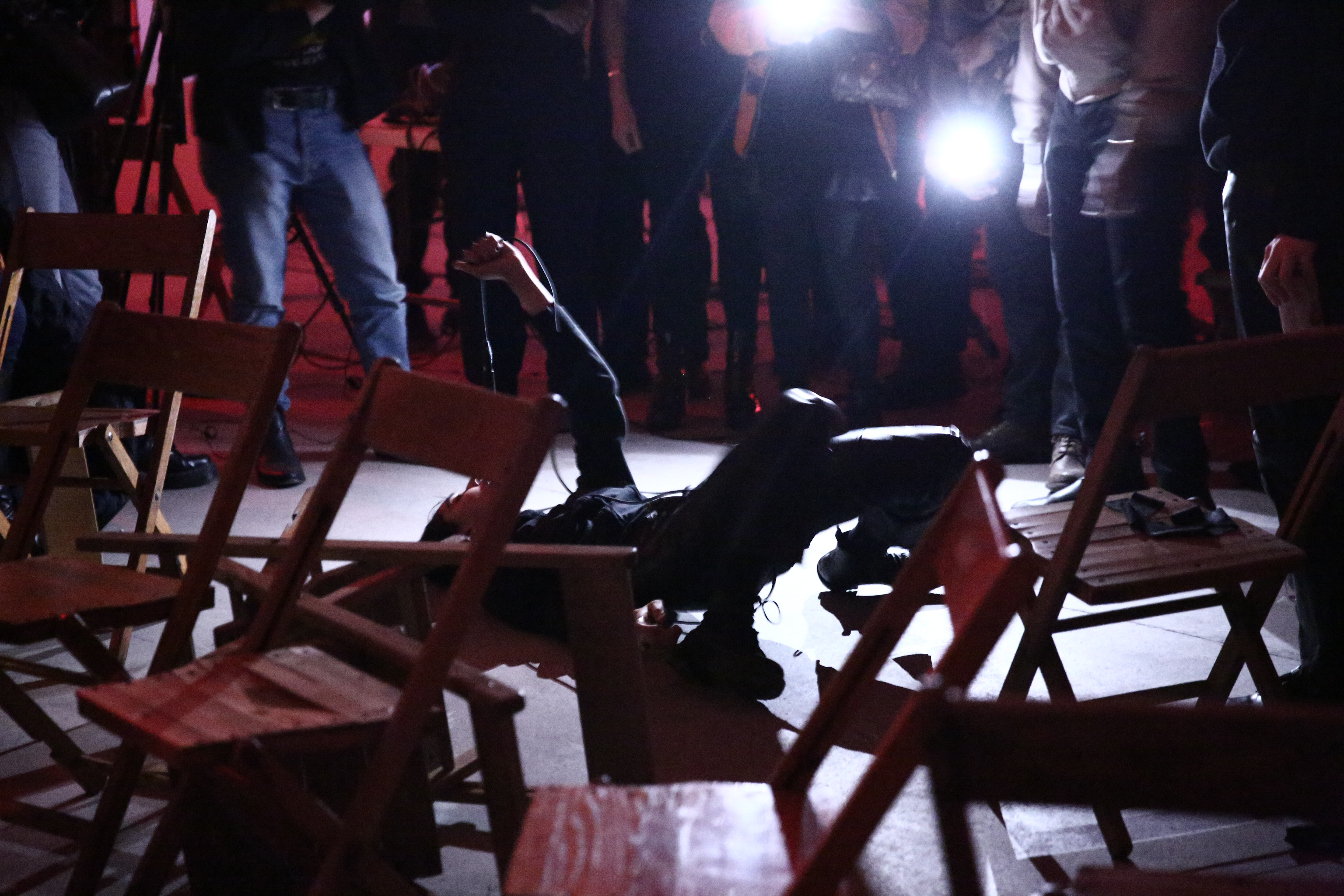 A figure wearing all black lies on their back on a concrete floor. Their hips and knees are lifted off the floor, with their hand holding a dangling microphone by the cord. There is a crowd of people arranged in a semi-circle behind the performer, and disordered empty chairs in the foreground.