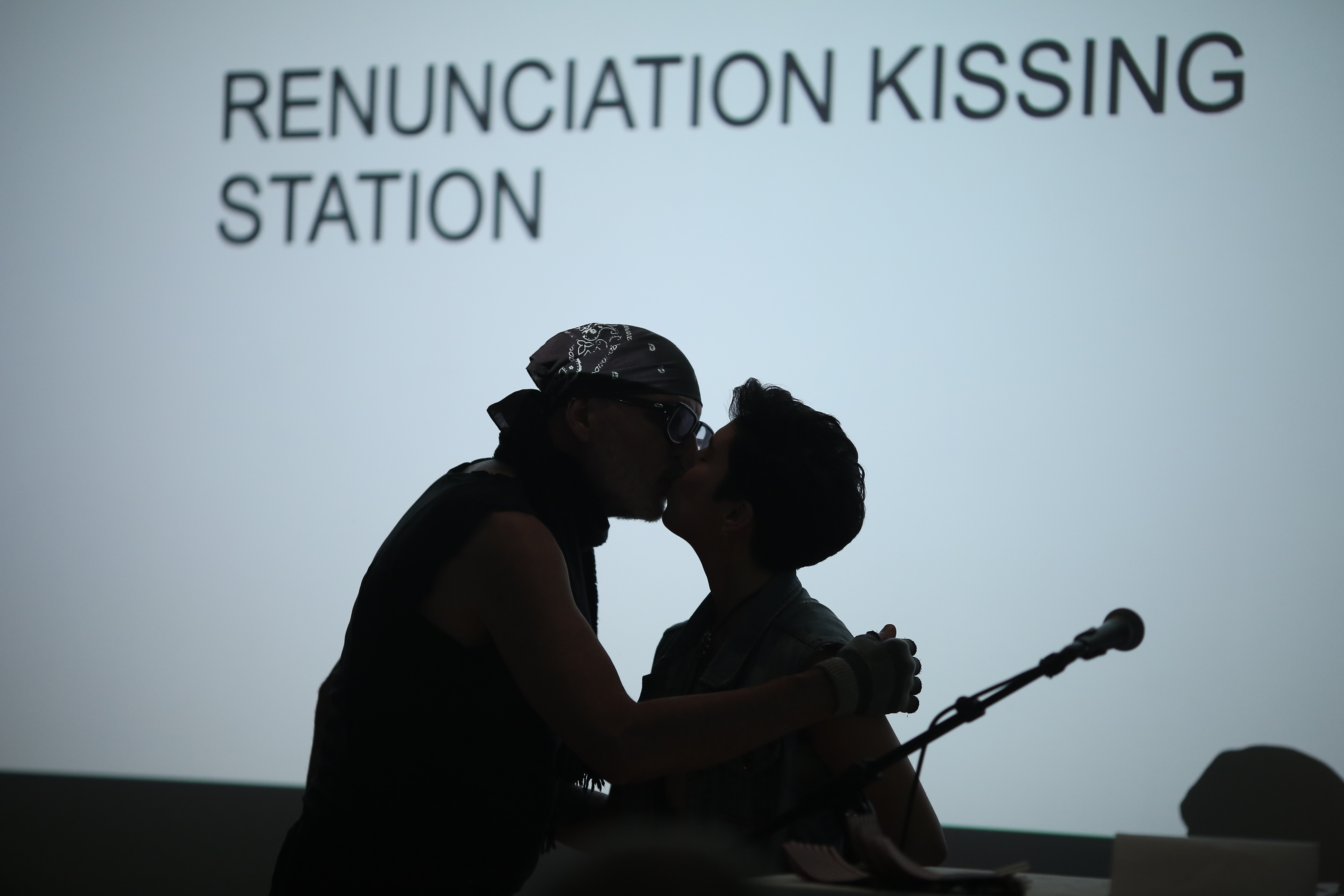 Two silhouetted figures kissing behind a microphone. They are backlit by a white projection with black capitalized text reading 