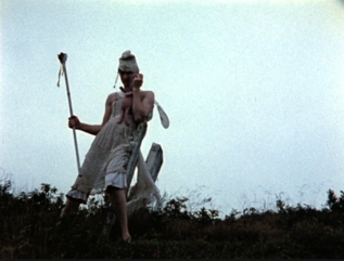Photo of a person in a white robe and headdress carrying a pole in their right hand, standing outdoors against a grey sky