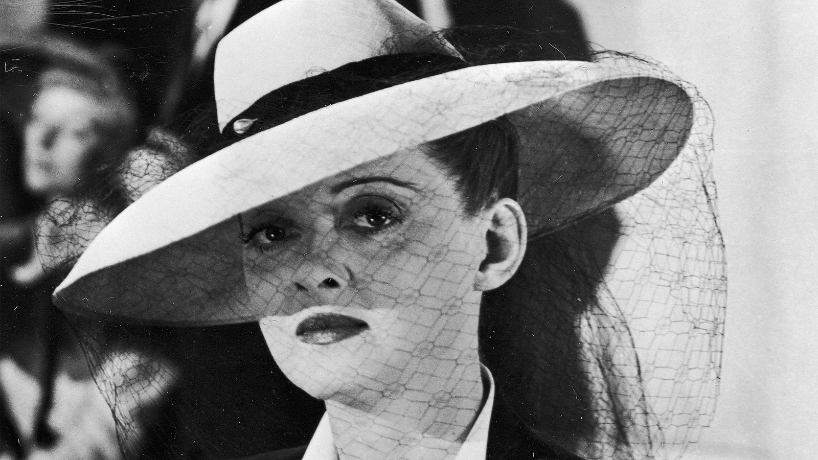 A black and white image of a woman looking into the camera. She has a wide-brimmed hat with a sheer veil over her face, and dark makeup.