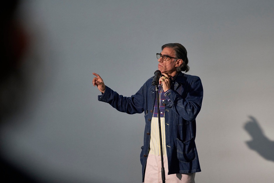 A figure stands behind a microphone stand, one hand raised to their chin and the other arm up and pointing away from themselves. The figure stands in front of a blank background.