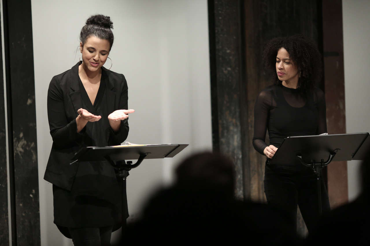 Two figures in dark clothing stand before two music stands in front of a seated audience. The figure on the right looks towards the figure on the left, who looks down at their two hands. Their hands are turned, palms up, towards the sky, as if miming holding something.