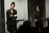 Two figures in dark clothing stand before two music stands in front of a seated audience. The figure on the right looks towards the figure on the left, who looks down at their two hands. Their hands are turned, palms up, towards the sky, as if miming holding something.