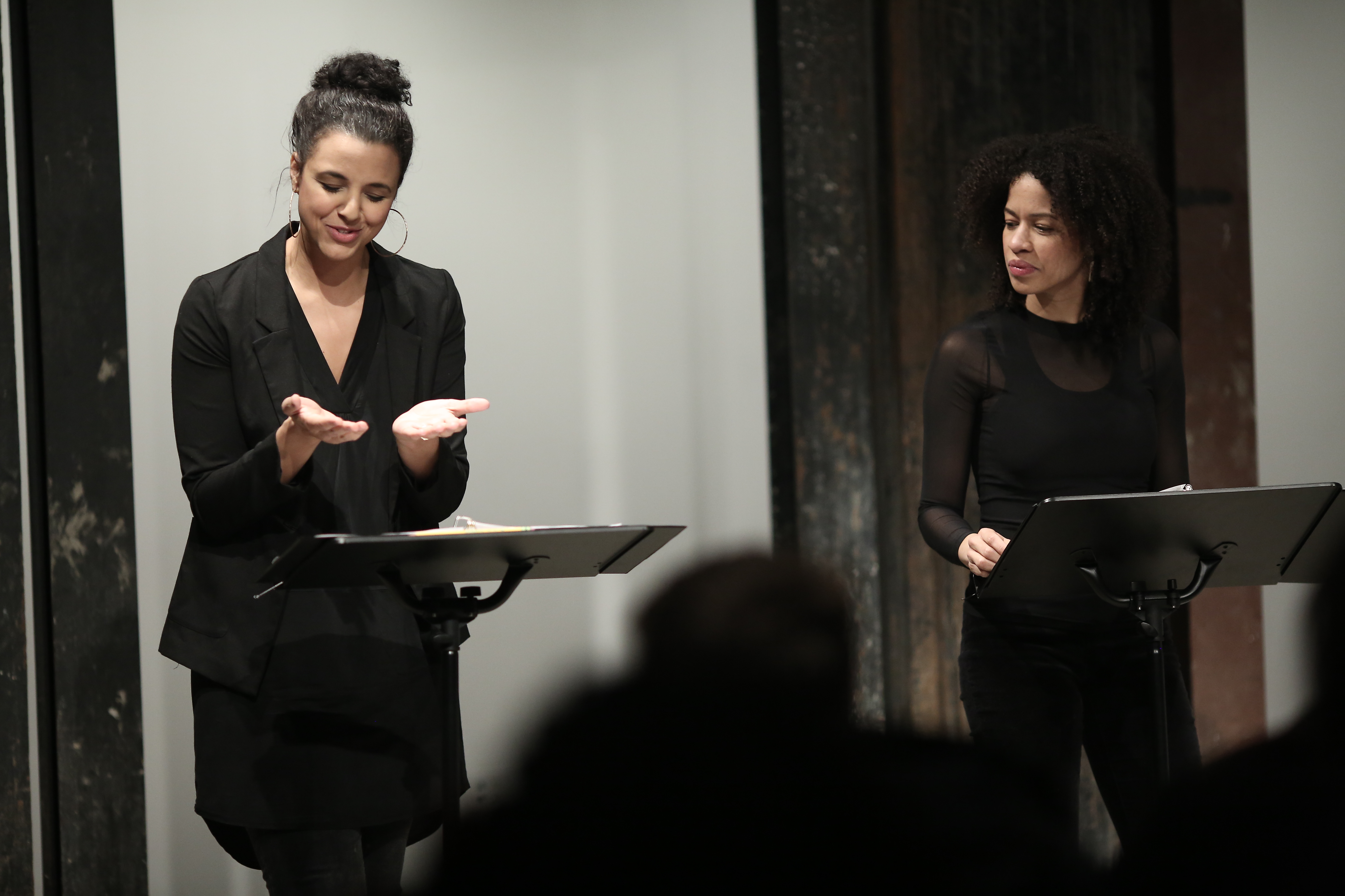 Two figures in dark clothing stand before two music stands in front of a seated audience. The figure on the right looks towards the figure on the left, who looks down at their two hands. Their hands are turned, palms up, towards the sky, as if miming holding something.