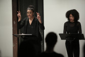 Two figures in dark clothing stand before two music stands in front of a seated audience. The figure on the left speaks, raising up both hands by her face, her fingers making 
