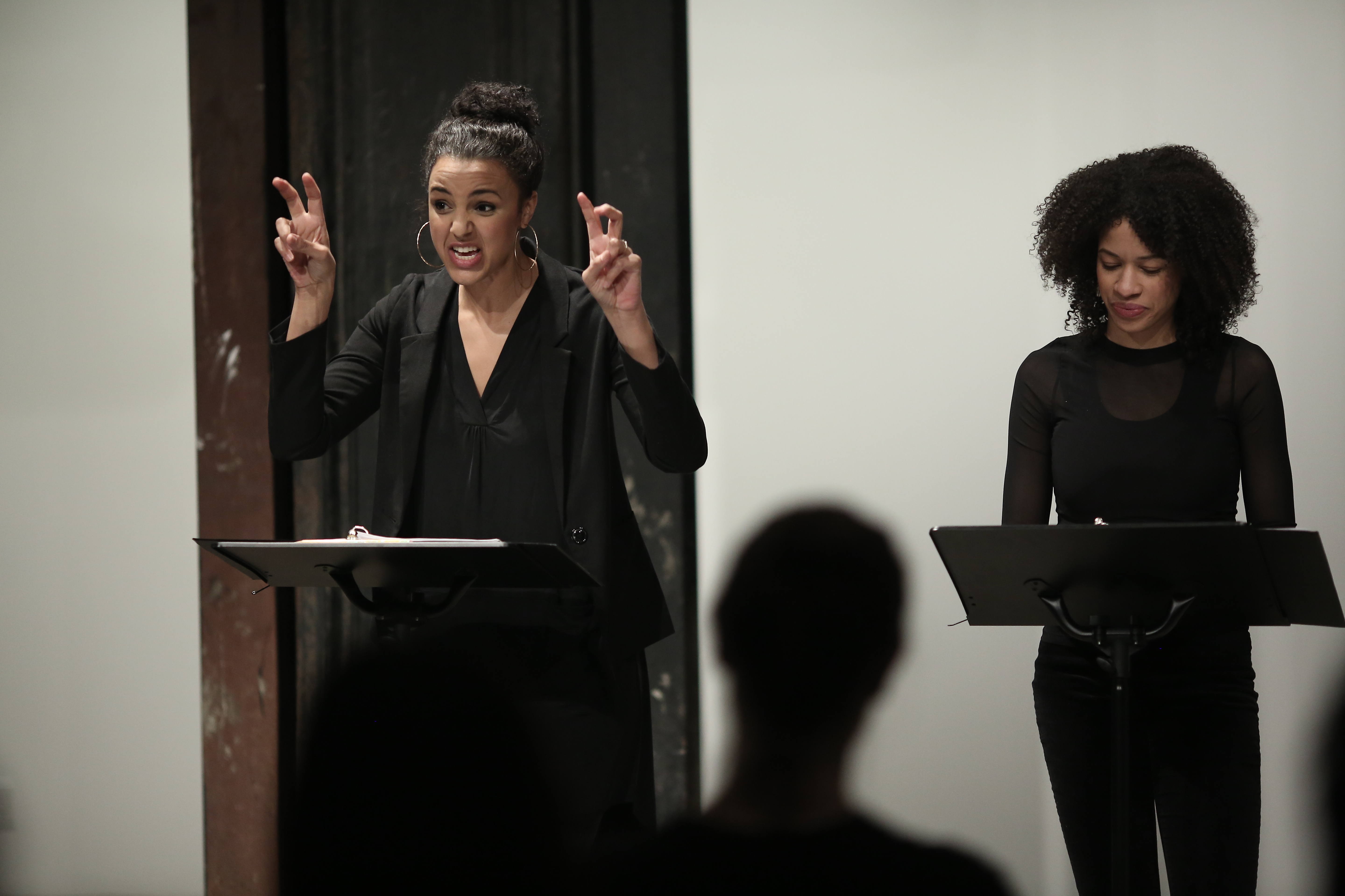 Two figures in dark clothing stand before two music stands in front of a seated audience. The figure on the left speaks, raising up both hands by her face, her fingers making 