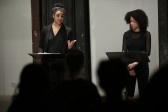 Two figures in dark clothing stand before two music stands in front of a seated audience. The figure on the right appears to read from a text propped on the stand, while the figure on the left looks towards the audience, speaking and gesturing towards them with one hand.