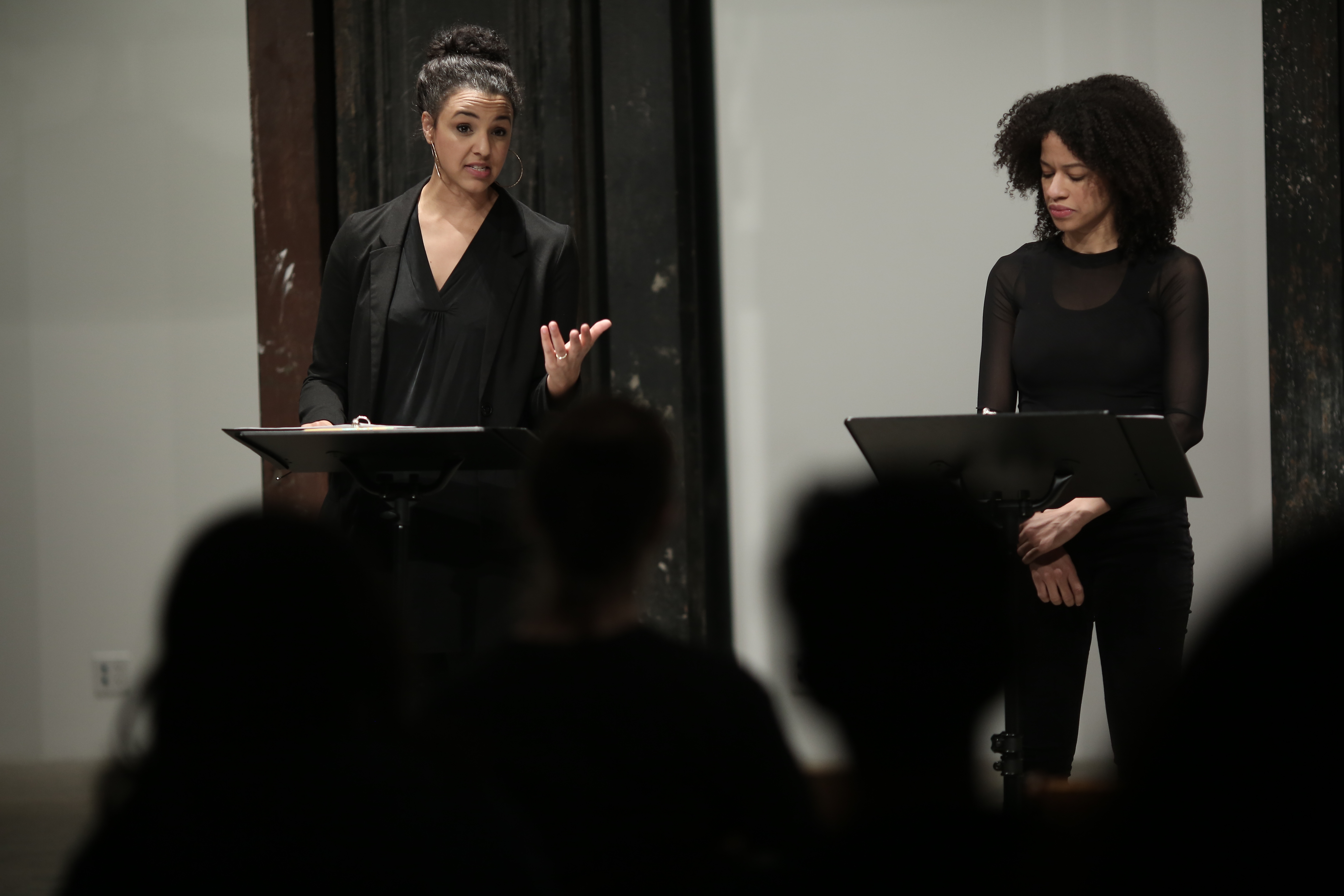 Two figures in dark clothing stand before two music stands in front of a seated audience. The figure on the right appears to read from a text propped on the stand, while the figure on the left looks towards the audience, speaking and gesturing towards them with one hand.