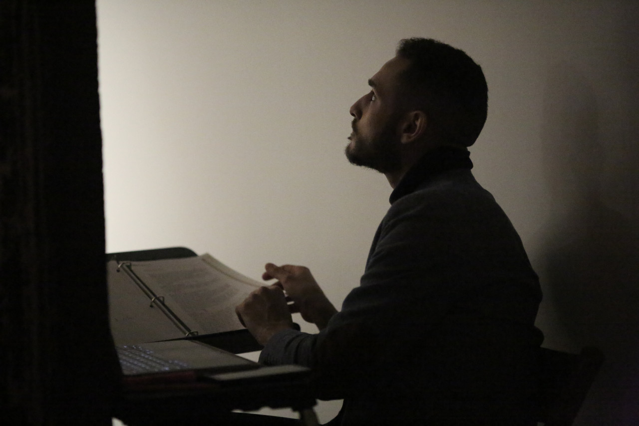 A figure sits in a dimly lit corner in profile, gazing forward. They flip through pages of a script displayed in front of them. An open laptop sits to their left.