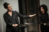 Two figures in dark clothing stand before two music stands. The figure on the right speaks and gazes forward. One of their hands rests on the music stand, the other reaches out in front of themselves, their fingers extended.