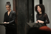 Two figures in dark clothing stand before two music stands in front of a seated audience. The figure on the right is speaking, her hand pinched in a small gesture at her torso.