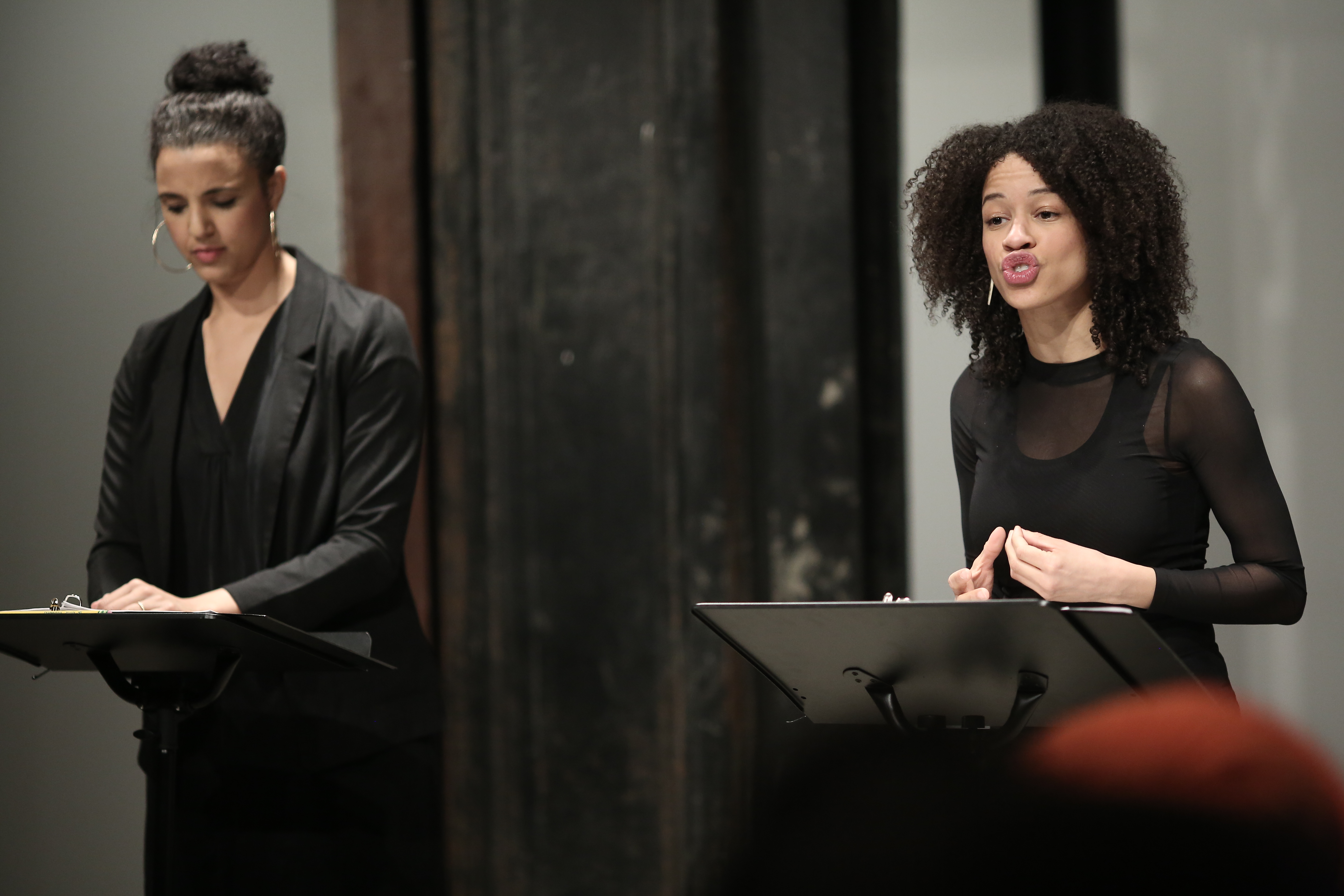 Two figures in dark clothing stand before two music stands in front of a seated audience. The figure on the right is speaking, her hand pinched in a small gesture at her torso.