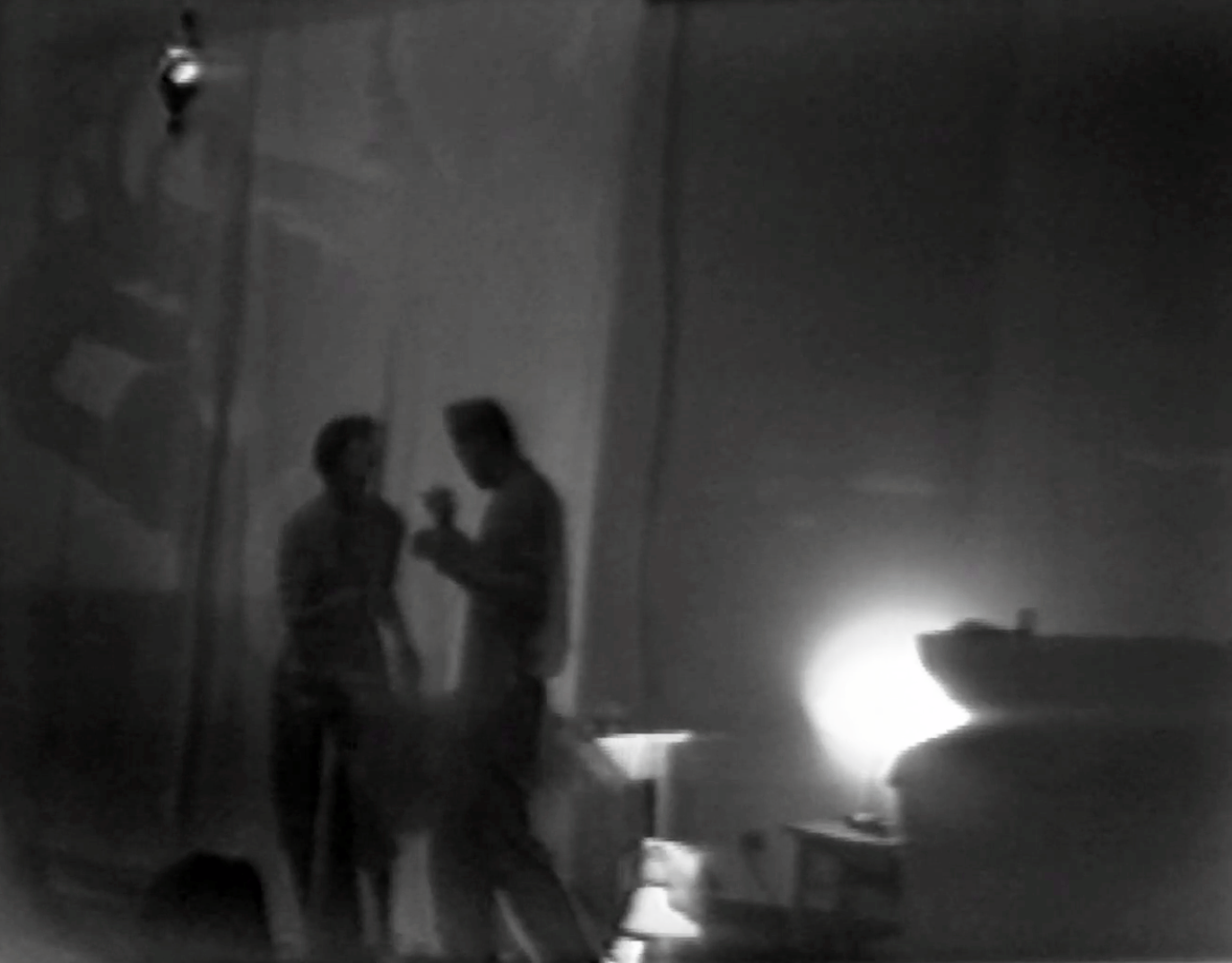 A black and white image of two blurry figures talking to each other in a dark room.