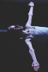A girl lays on a dark background. She wears a white leotard. Her arms are stretched straight out on either side, with her palms facing upwards. She gazes upwards, illuminated by a soft light.