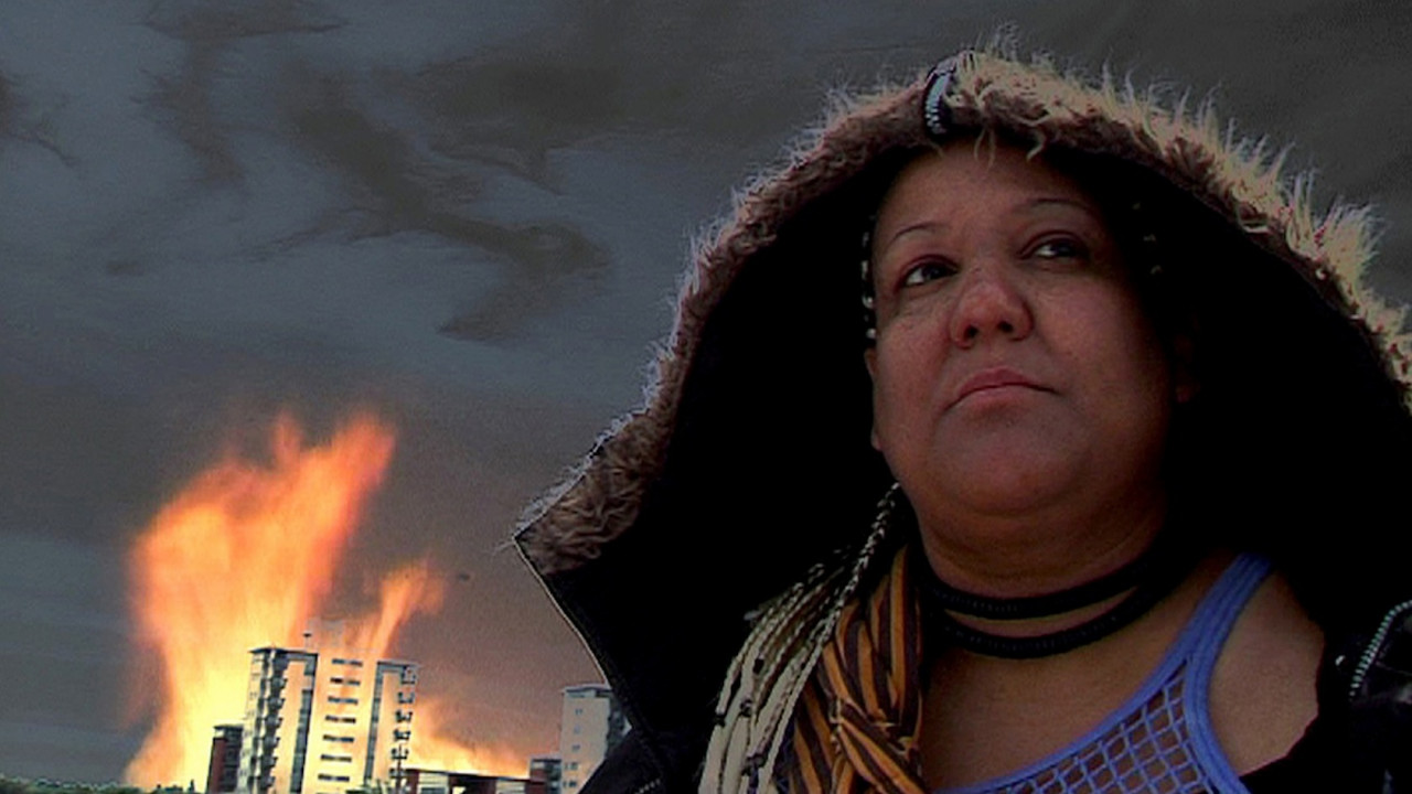 A woman with a hood looks diagonally upward, away from the camera. Behind her, a large building is on fire and the sky is dark.