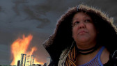 A woman with a hood looks diagonally upward, away from the camera. Behind her, a large building is on fire and the sky is dark.