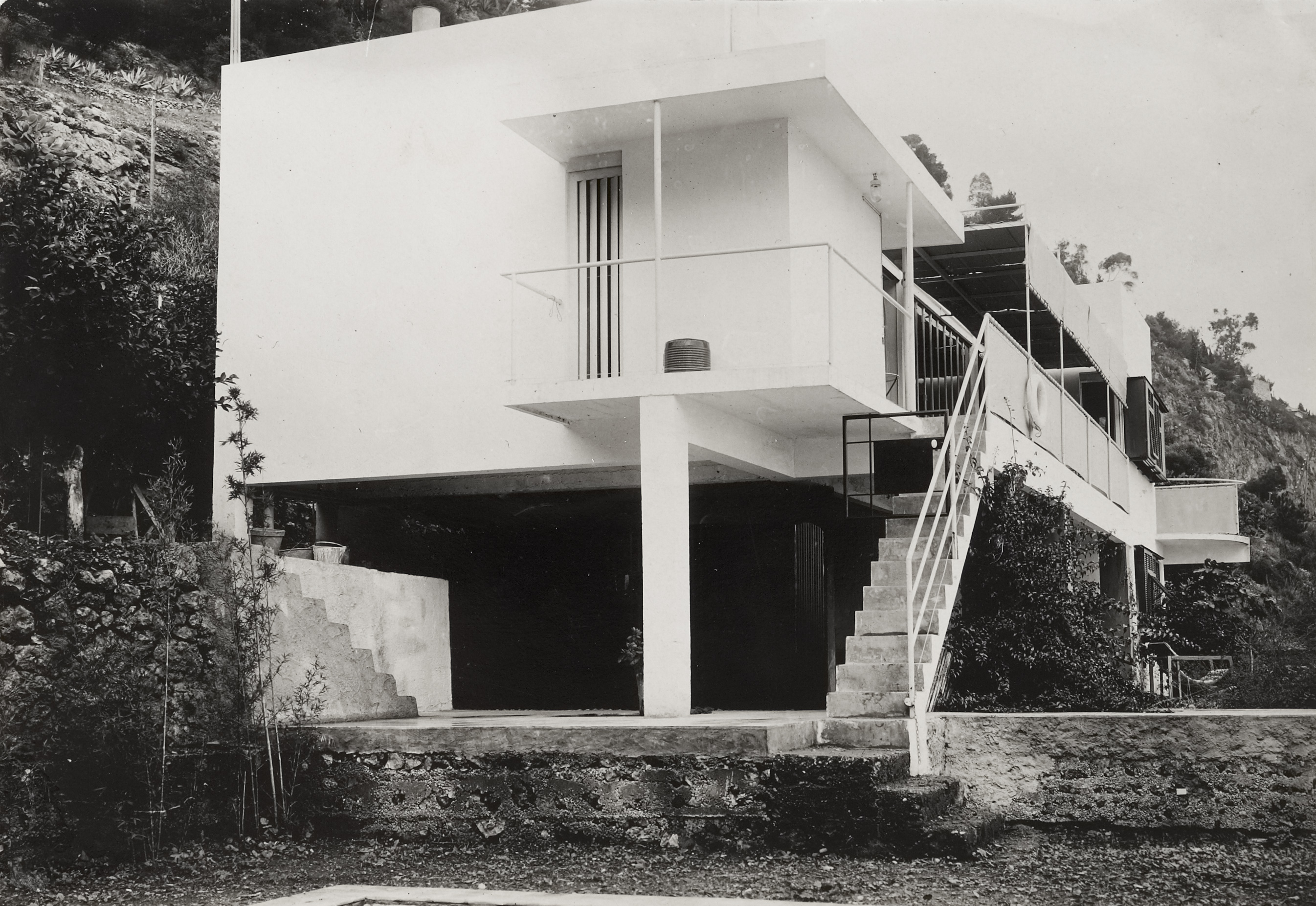 A black and white image of the exterior of a modern house standing on stilts.