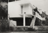A black and white image of the exterior of a modern house standing on stilts.