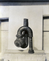 A black pipe coming out of a concrete slab beneath it, twisting around, pointed up and cut off, against a whitish slab behind it, as if to frame the pipe.