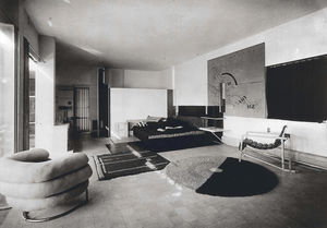 Black-and-white photo of the interior of a house, with several paintings, a round and square rug, and two armchairs, of various styles considered modern around the early 20th century.