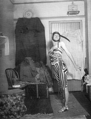 A black-and-white image of a woman wearing a striped dress and clip-on hat, eyes closed, arms stretched behind her. She stands in a room with several linens pinned to the walls and floor