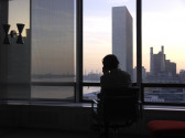 A figure sits in a chair facing—and silhouetted by—a soft, sunset-bathed city skyline and river. Their back faces the viewer, and they appear to rest their head on their hand. The space around them resembles a corporate office space.